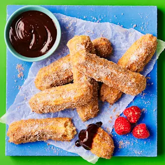 Sugar coated churros dipped in chocolate sauce on parchment paper on blue board with green background. Three pink raspberries on the side. 