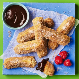 Sugar coated churros dipped in chocolate sauce on parchment paper on blue board with green background. Three pink raspberries on the side. 