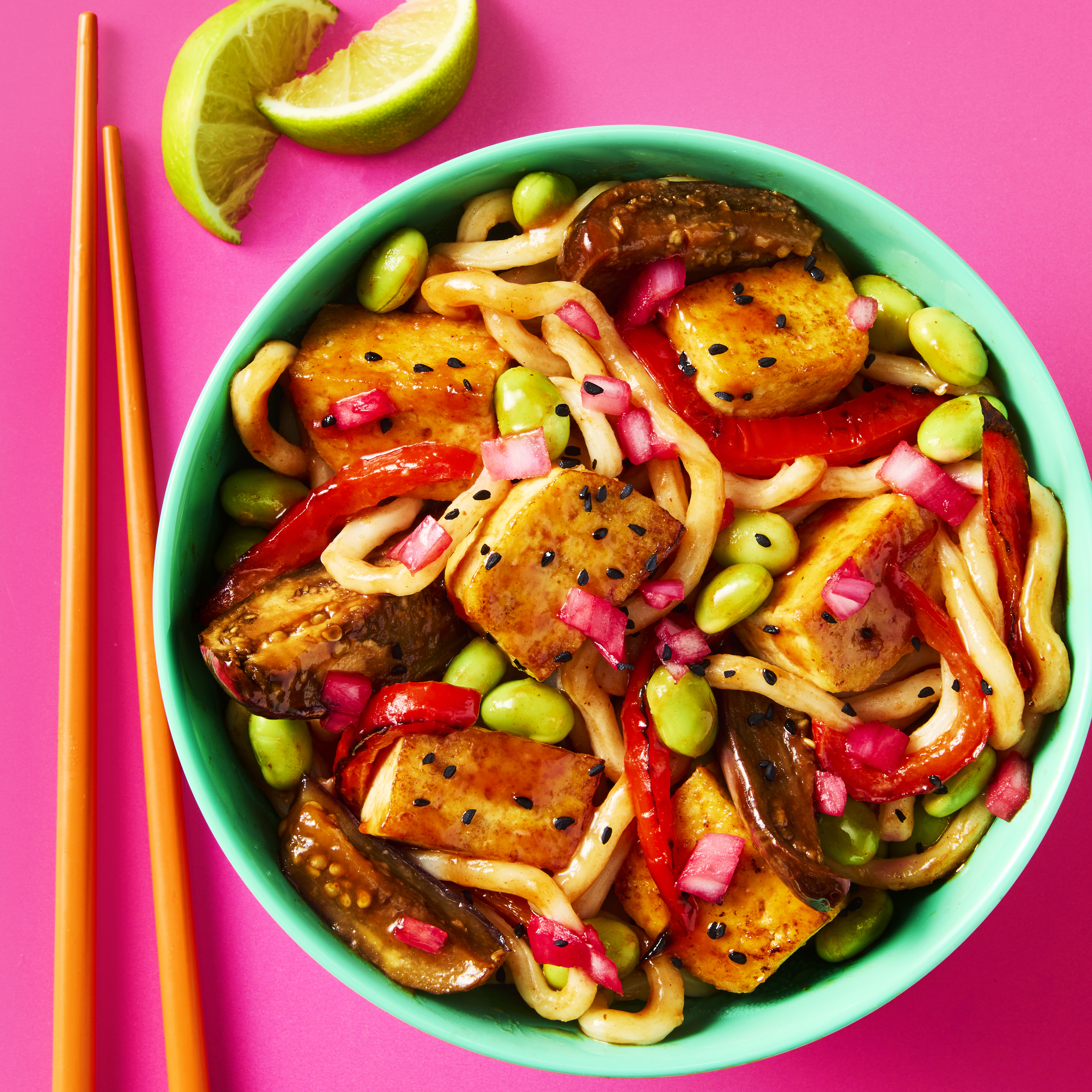 Tofu & Aubergine Miso Yaki Udon served in a bowl with chopsticks and a wedge of lime on the side.