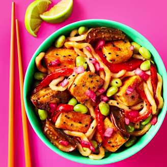 Tofu & Aubergine Miso Yaki Udon served in a bowl with chopsticks and a wedge of lime on the side.