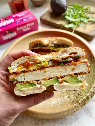 Hand holding bagel, with crispy tofu, avocado, pink Cauldron tofu packaging in background, greens on wooden chopping board.