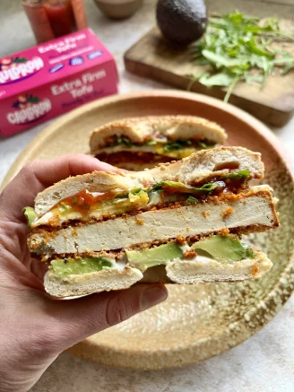 Hand holding bagel, with crispy tofu, avocado, pink Cauldron tofu packaging in background, greens on wooden chopping board.