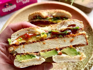 Hand holding bagel, with crispy tofu, avocado, pink Cauldron tofu packaging in background, greens on wooden chopping board.