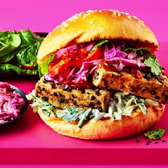 Marinated tofu slices in a brioche burger bun with salad, tomatoes and pink Indian slaw on a pink board.
