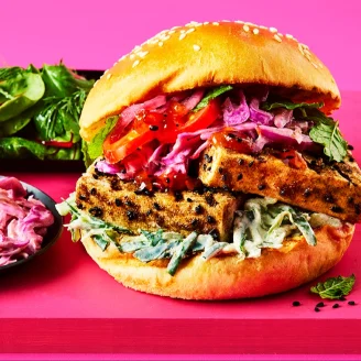 Marinated tofu slices in a brioche burger bun with salad, tomatoes and pink Indian slaw on a pink board.