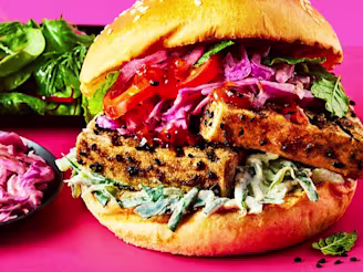 Marinated tofu slices in a brioche burger bun with salad, tomatoes and pink Indian slaw on a pink board.
