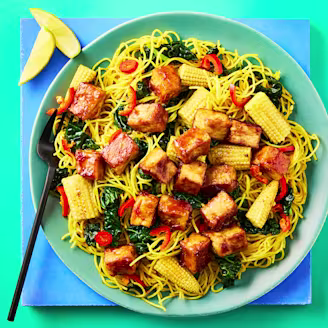 Cauldron Tofu Pieces in a soy and garlic sauce with Singapore noodles and chopped vegetables on a green plate with black cutlery. 