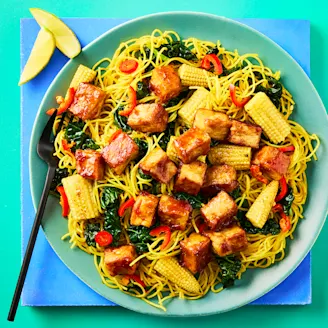 Cauldron Tofu Pieces in a soy and garlic sauce with Singapore noodles and chopped vegetables on a green plate with black cutlery. 