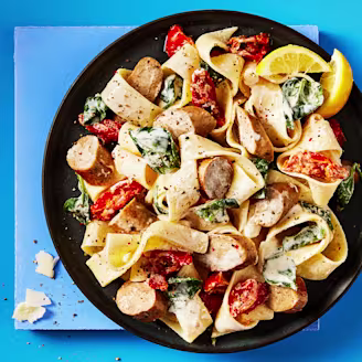 Cauldron Cumberland sausages and pasta with vegetables served in a dark dish with lemon slices on the side, the background is blue