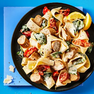 Cauldron Cumberland sausages and pasta with vegetables served in a dark dish with lemon slices on the side, the background is blue