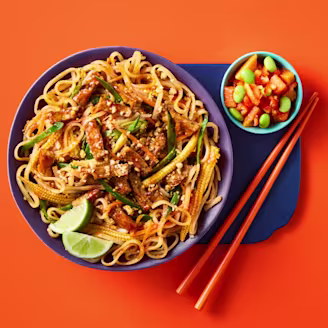 Cauldron Marinated Tofu with noodles and Pad Thai sauce with baby corn, bean sprouts, mange tout and lime wedges to garnish. A side of papaya and edamame salad is in a light blue bowl to the side with orange chopsticks and an orange background.
