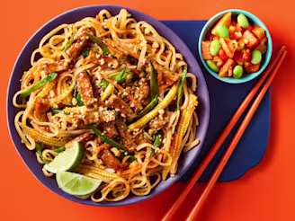 Cauldron Marinated Tofu with noodles and Pad Thai sauce with baby corn, bean sprouts, mange tout and lime wedges to garnish. A side of papaya and edamame salad is in a light blue bowl to the side with orange chopsticks and an orange background.