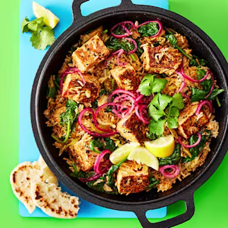 Biryani made using Cauldron Tofu topped with red onion, coriander and lemon slices served in a large dark pot with a green background 