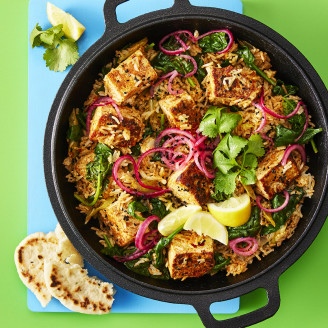 Biryani made using Cauldron Tofu topped with red onion, coriander and lemon slices served in a large dark pot with a green background 