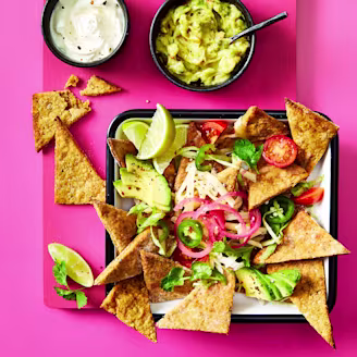 Nachos made using Cauldron Falafel served with avocado, tomatoes, red pickled onions, limes and dips on the side. Served in a tray with a pink background. 