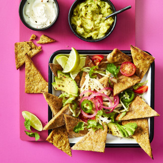 Nachos made using Cauldron Falafel served with avocado, tomatoes, red pickled onions, limes and dips on the side. Served in a tray with a pink background. 