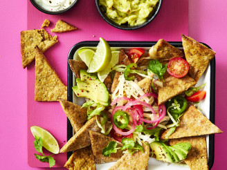 Nachos made using Cauldron Falafel served with avocado, tomatoes, red pickled onions, limes and dips on the side. Served in a tray with a pink background. 