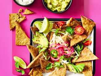 Nachos made using Cauldron Falafel served with avocado, tomatoes, red pickled onions, limes and dips on the side. Served in a tray with a pink background.