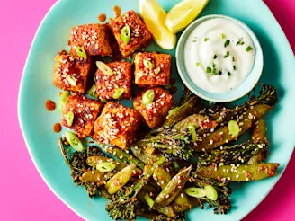 Tofu in buffalo sauce on blue plate with tenderstem broccoli, ranch dipping sauce and lemon wedges, on pink background.