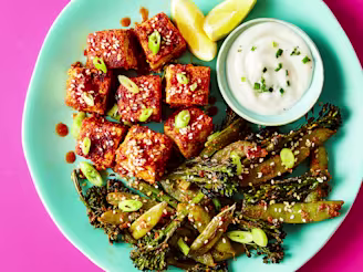Tofu in buffalo sauce on blue plate with tenderstem broccoli, ranch dipping sauce and lemon wedges, on pink background.