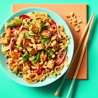 Rice noodles in a blue bowl topped with Cauldron Tofu Pieces, green and red vegetables with chopsticks on an orange placemat. 