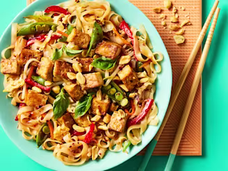 Rice noodles in a blue bowl topped with Cauldron Tofu Pieces, green and red vegetables with chopsticks on an orange placemat.