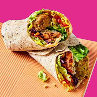 A burrito cut open to showcase the filling including Cauldron spiced falafel, rice and veggies served on an orange mat with a pink background.