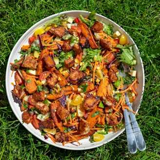 A serving bowl full of BBQ Tofu Rainbow Summer Salad, a spoon and fork on a grassed background