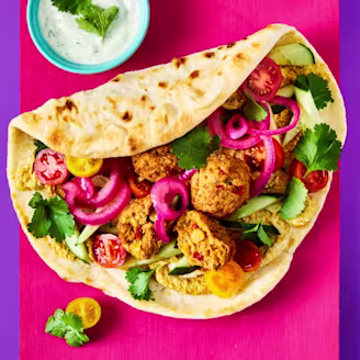 Pita with Cauldron Butternut and Red Chilli bites, chopped cherry tomatoes, coriander, pickled red onion and a side dish of yoghurt dip on a vibrant pink and purple background. 
