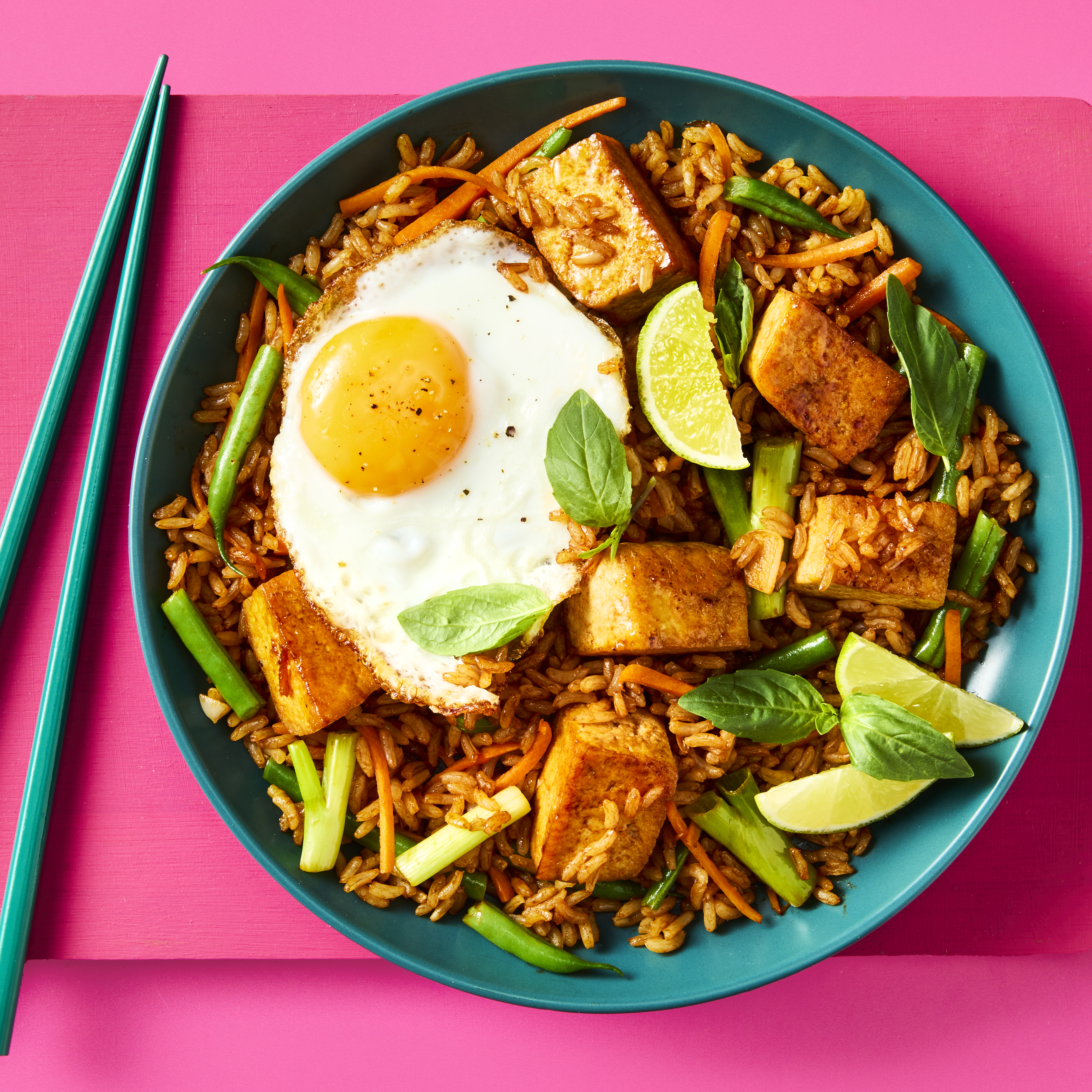Nasi Goreng on a plate with golden tofu, a fried egg, lime wedges, Thai basil, and chopsticks on the side.
