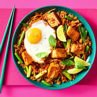 Nasi Goreng on a plate with golden tofu, a fried egg, lime wedges, Thai basil, and chopsticks on the side.
