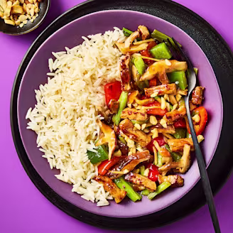 Oyster sauce stir fry served in a purple bowl with white rice, chopped vegetables and a fork with a side of nuts to garnish.