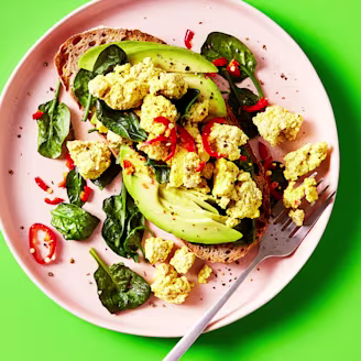 Tofu scramble served with spinach and avocado on sourdough bread topped with chilli on a pink plate with a green background