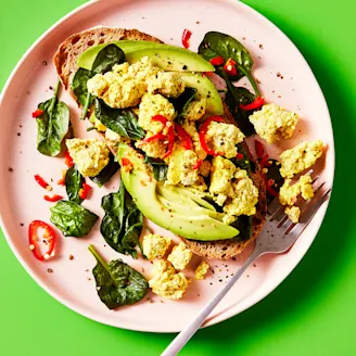 Tofu scramble served with spinach and avocado on sourdough bread topped with chilli on a pink plate with a green background