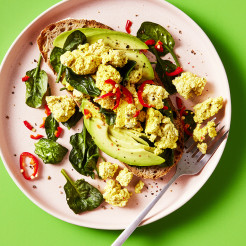 Tofu scramble served with spinach and avocado on sourdough bread topped with chilli on a pink plate with a green background