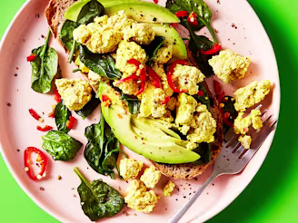 Tofu scramble served with spinach and avocado on sourdough bread topped with chilli on a pink plate with a green background