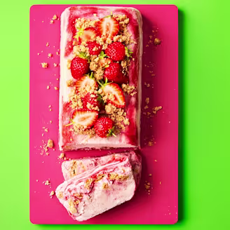Strawberries and crumbled biscuit on top of rectangular shaped semifreddo desert, sliced at the end on a pink board with green background.