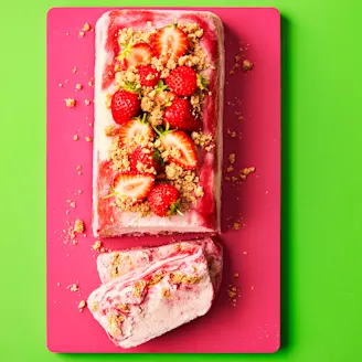 Strawberries and crumbled biscuit on top of rectangular shaped semifreddo desert, sliced at the end on a pink board with green background.