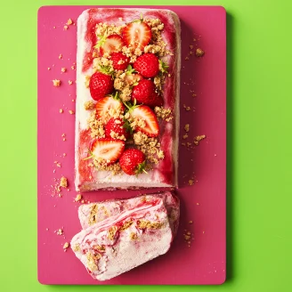 Strawberries and crumbled biscuit on top of rectangular shaped semifreddo desert, sliced at the end on a pink board with green background.