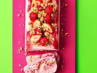 Strawberries and crumbled biscuit on top of rectangular shaped semifreddo desert, sliced at the end on a pink board with green background.