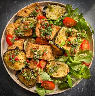 Beige plate on black table with tofu, aubergine, chopped tomatoes and greens with red and green chimichurri sauce and garnish