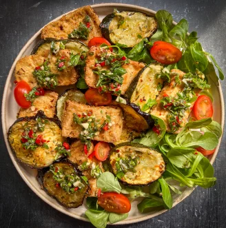 Beige plate on black table with tofu, aubergine, chopped tomatoes and greens with red and green chimichurri sauce and garnish