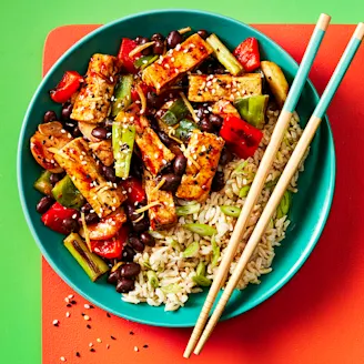 Black bean tofu with vegetables and a side of rice served in a blue dish with chopsticks on the side, the bowl is sitting on a red mat and the background is green. 