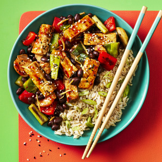 Black bean tofu with vegetables and a side of rice served in a blue dish with chopsticks on the side, the bowl is sitting on a red mat and the background is green. 