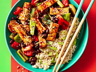 Black bean tofu with vegetables and a side of rice served in a blue dish with chopsticks on the side, the bowl is sitting on a red mat and the background is green.