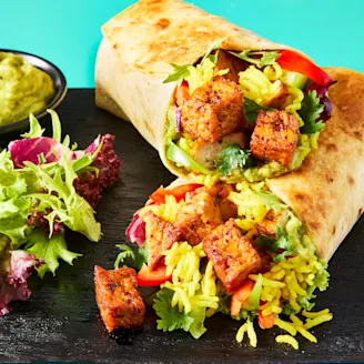 A Mexican Burrito made with Cauldron Tofu Pieces on a black serving board with a side salad and avocado dip.