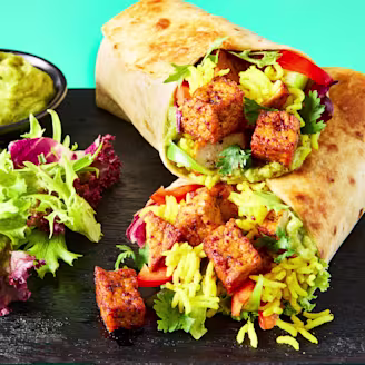 A Mexican Burrito made with Cauldron Tofu Pieces on a black serving board with a side salad and avocado dip.