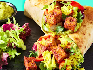 A Mexican Burrito made with Cauldron Tofu Pieces on a black serving board with a side salad and avocado dip.