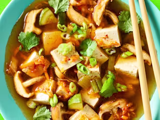 A Sundubu Jiggae with Cauldron Authentic Tofu and onions, garlic, kimchi, mushrooms, soy sauce and coriander leaves in a blue bowl on a green background.