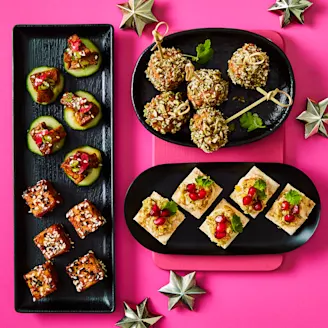 Assortment of vegan christmas canapes on black serving plates on a bright pink background.